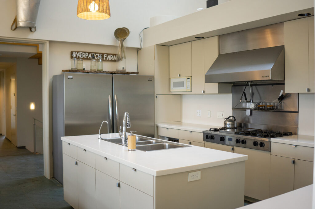 A view of the kitchem with a large, stainless steel refrigerator, stove and triple sink.